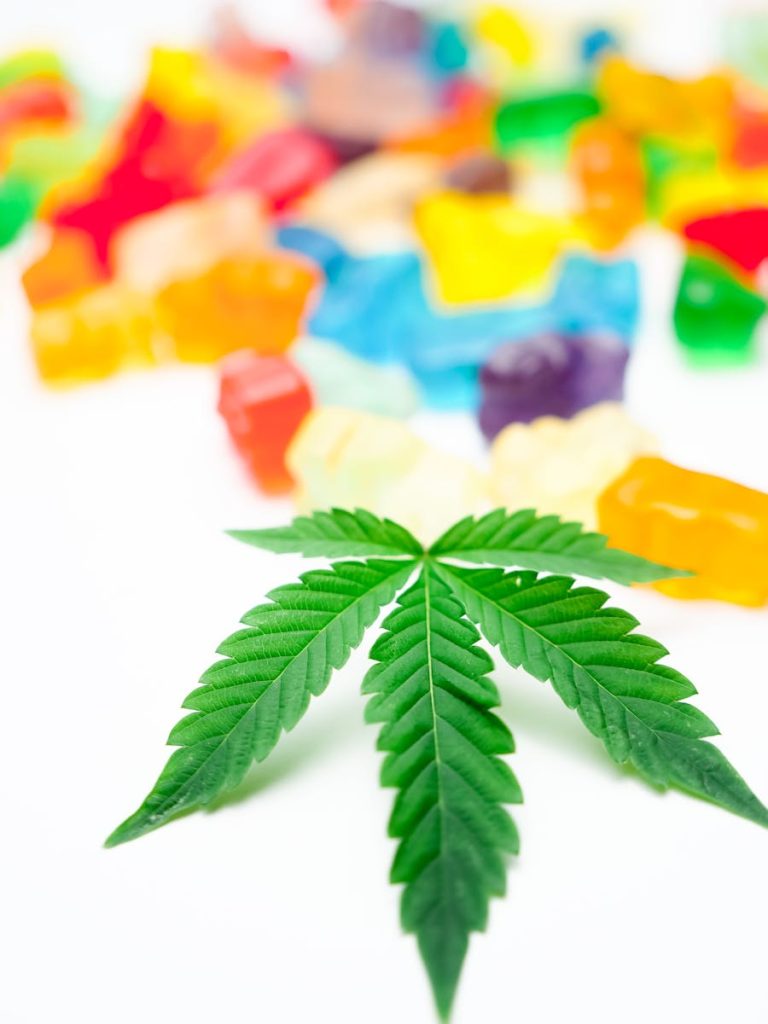 Close-up view of a cannabis leaf with an assortment of colorful gummy candies on a white background.